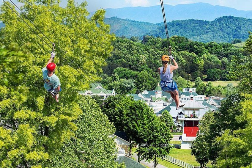 To personer svæver i zipline over et skovområde med udsigt til huse og bjerge i baggrunden. 1