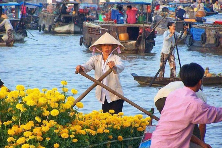 A river scene with boats, people in traditional attire, and yellow flowers in the foreground. 1