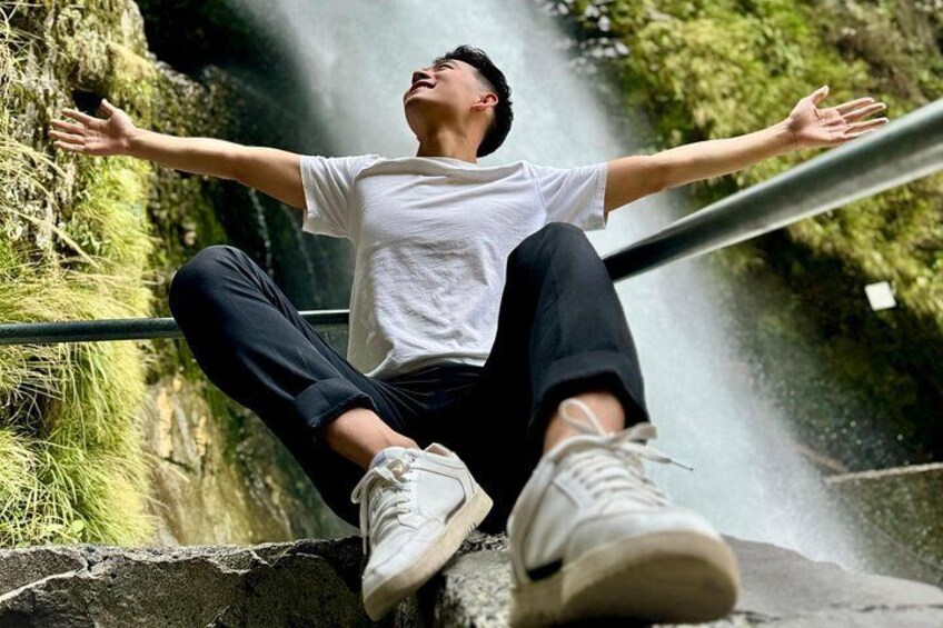 Una persona seduta su una sporgenza, con le braccia aperte, indossa una t-shirt bianca e pantaloni neri, con una strada e vegetazione sullo sfondo. 4