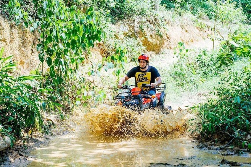A person riding a quad bike through a muddy stream surrounded by greenery. 3