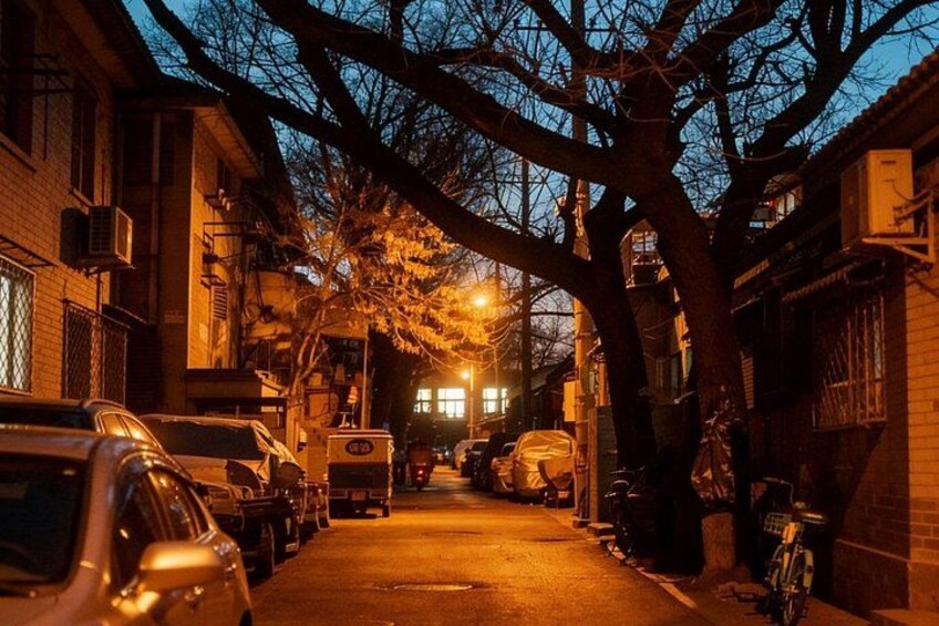 Una calle urbana estrecha al anochecer, con coches estacionados y edificios a ambos lados. 2