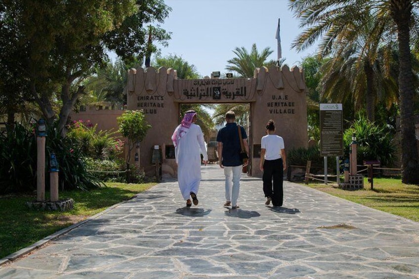 A pathway leading to the UAE Heritage Village entrance with palm trees and a clear sky. 1
