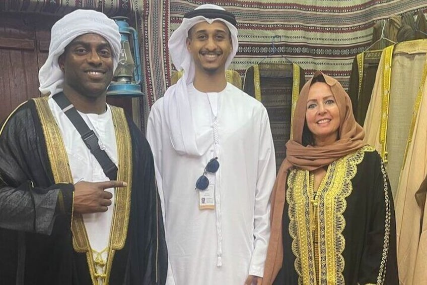Three people in traditional attire, standing in a room with patterned rugs and a wooden cabinet. 5