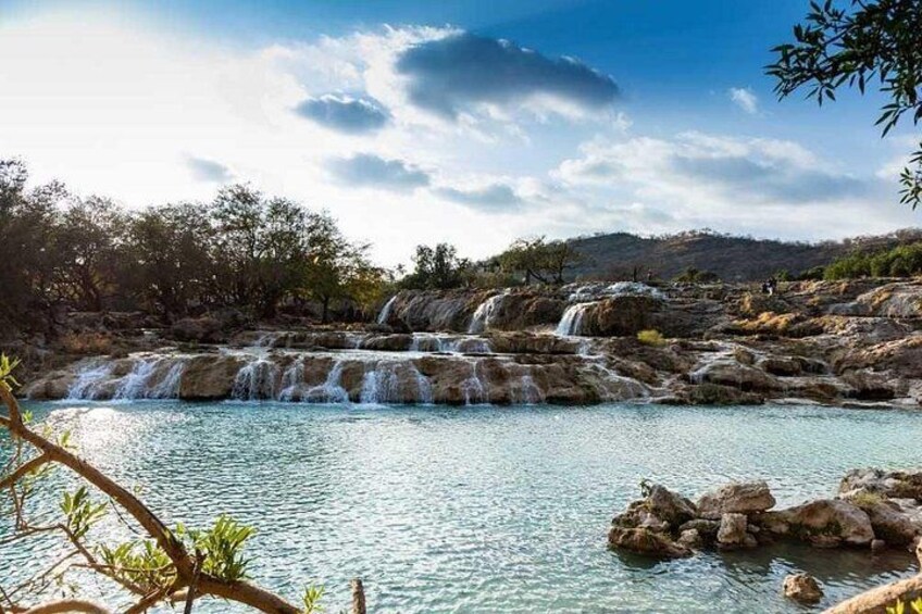 A waterfall cascading over rocks into a clear pool of water, surrounded by trees and hills. 2