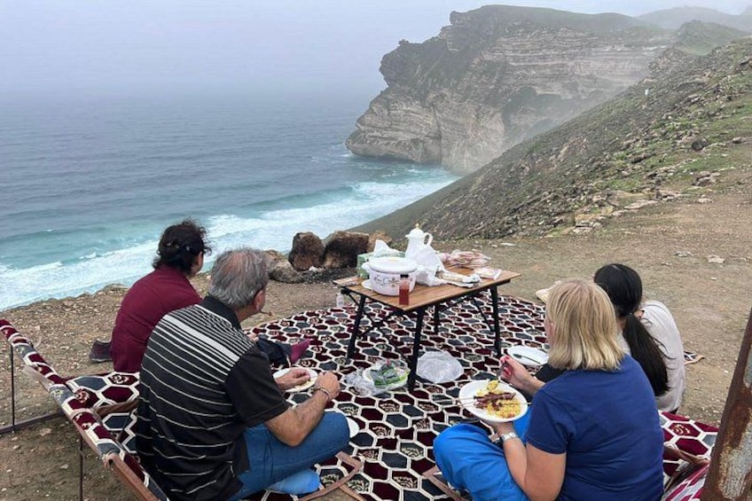 Eine Gruppe von Personen sitzt auf gemusterten Stühlen an einem Tisch auf einer Klippe mit Blick auf das Meer. 1