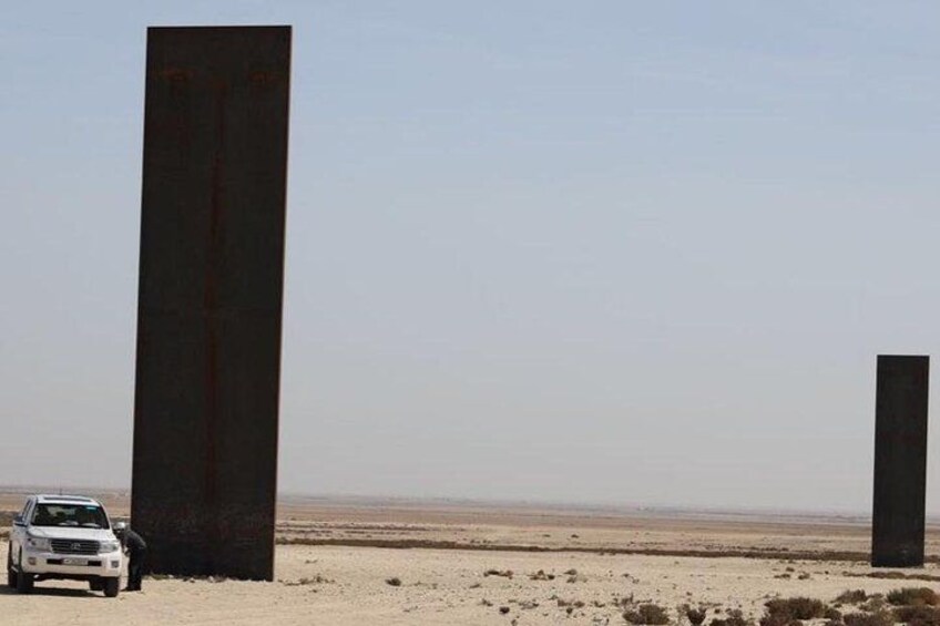 Paisaje desértico con dos estructuras verticales grandes y oscuras, y un vehículo blanco estacionado cerca. 5