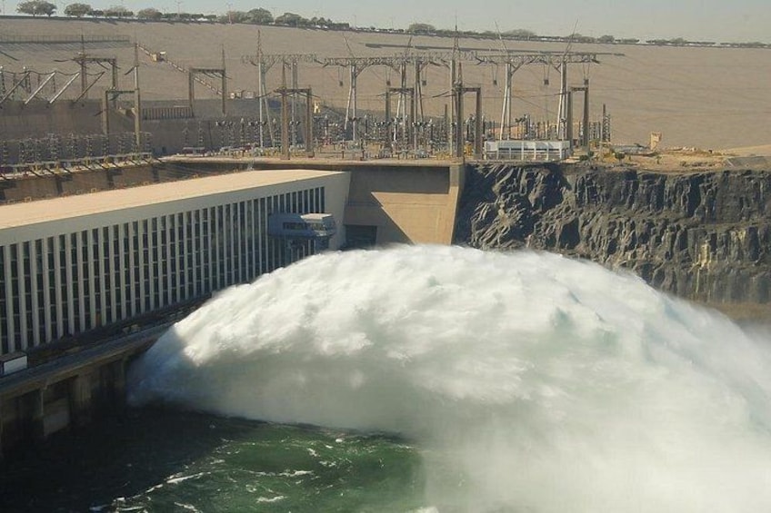 Een dam met water dat eroverheen stroomt, een controlegebouw en hoogspanningslijnen op de achtergrond. 2