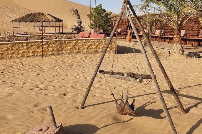 A desert landscape with a wooden swing set, a thatched-roof hut, and palm trees. 4