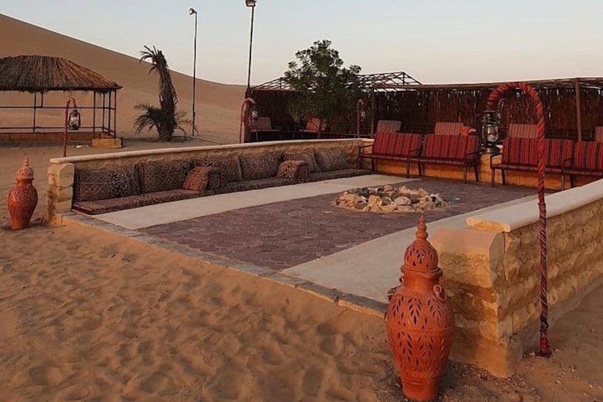 A desert landscape with a seating area, a thatched-roof structure, and a large vase. 5