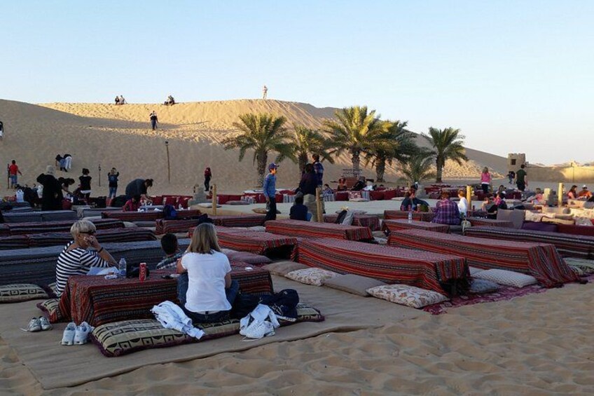 Un paysage désertique où l’on voit des personnes se détendre sur des tapis aux motifs colorés, des palmiers et, au loin, une dune de sable. 5