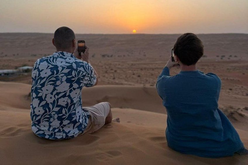 Dos personas sentadas en dunas de arena fotografiando la puesta de sol. 3