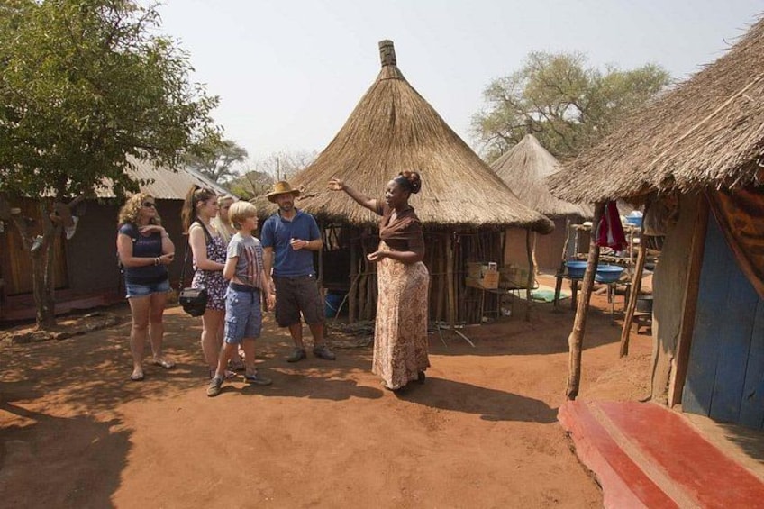 A group of people in a rural setting with thatched-roof huts and a dirt ground. 1