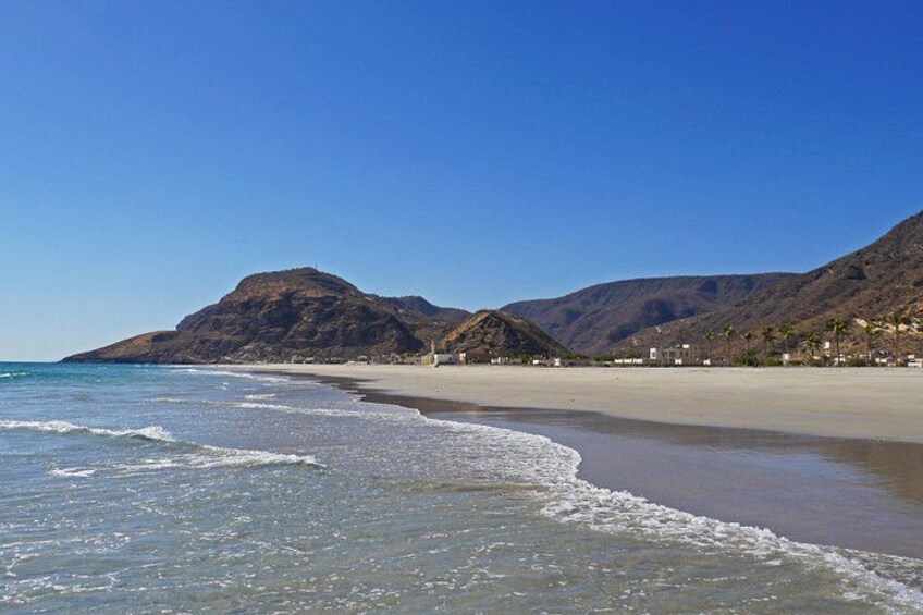 Een strand met helderblauw water, een zandstrand en bergen op de achtergrond. 3