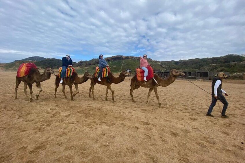 Four camels with riders in a sandy area, a person leading the camels, and a hilly landscape in the background. 5