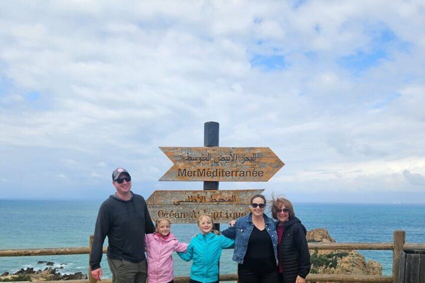 A family standing in front of a sign with directions in multiple languages, including Arabic and French, by the sea. 4