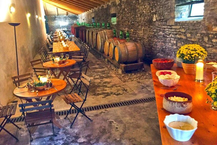 Una bodega rústica con mesas y sillas de madera, barriles y una mesa larga con comida y bebidas. 1