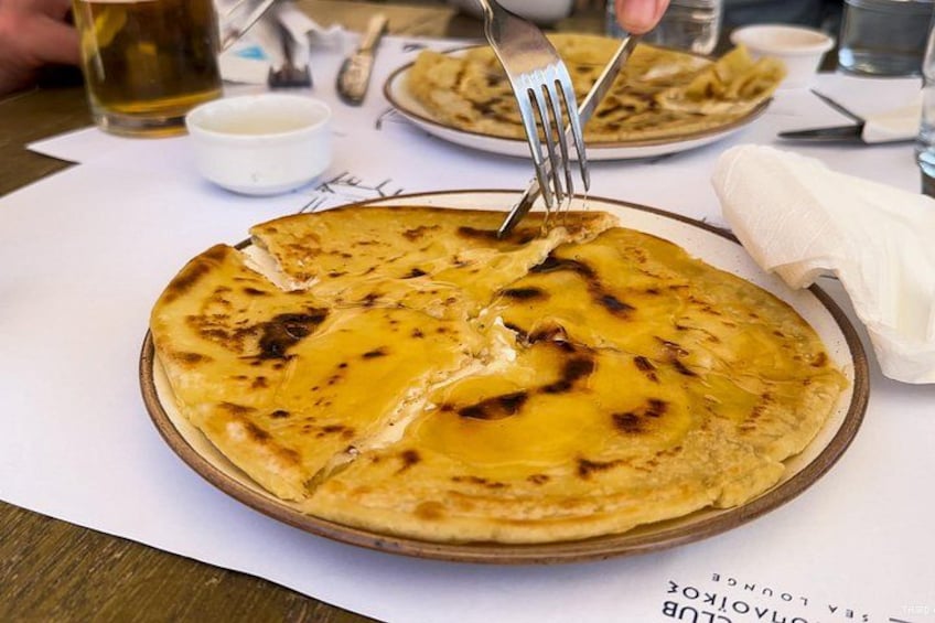 A round flatbread with a golden-brown crust, a fork cutting into it, and a small white bowl in the background. 2