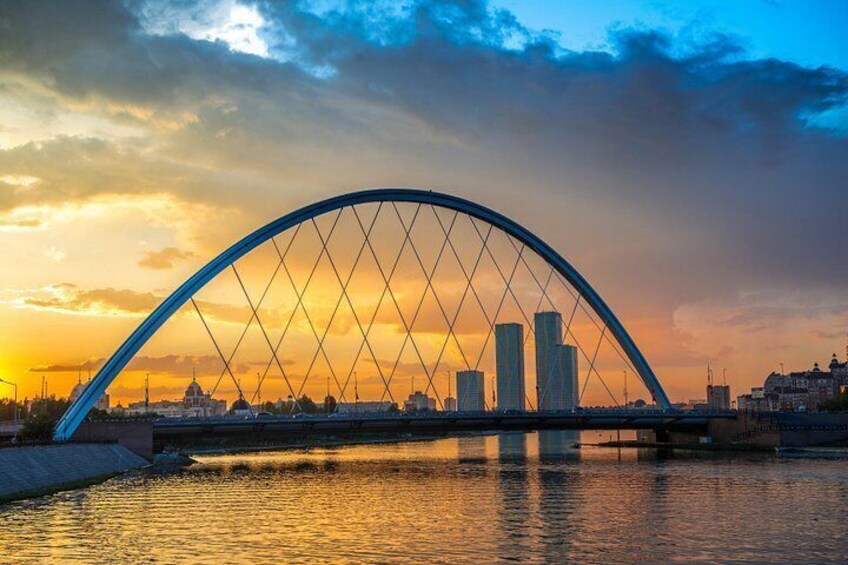 Eine große, gewölbte Brücke über einem Gewässer, im Hintergrund die Skyline einer Stadt bei Sonnenuntergang. 2