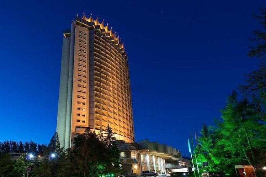 Ett högt hotell med en böjd fasad, upplyst av ett varmt ljus, omgivet av träd och en klar himmel. 5
