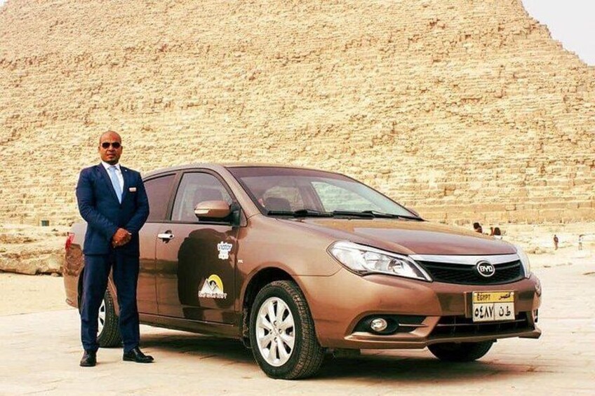 A man in a suit standing next to a brown GMC vehicle in front of a large pyramid. 2
