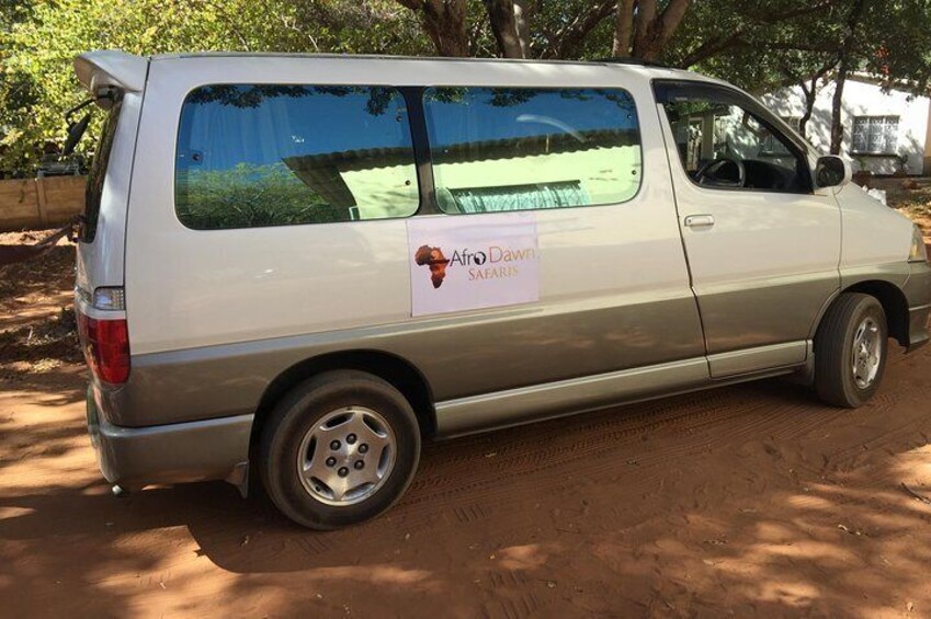 A silver van parked on a dirt road with a sign on the side that reads "Aero Dawn Safaris." 1