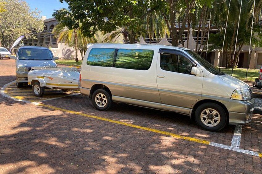 A parked silver minivan with a trailer in a parking lot with trees and buildings in the background. 3