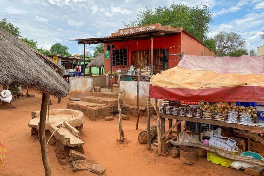 Escena de mercado rural con una tienda llamada 'IN GOD WE TRUST SHOP' y un puesto improvisado que vende productos. 5
