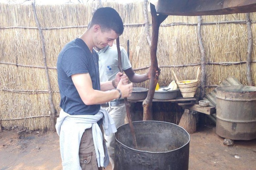 Una persona cocina al aire libre, con una olla grande, una mesa de madera y un muro de paja al fondo. 2