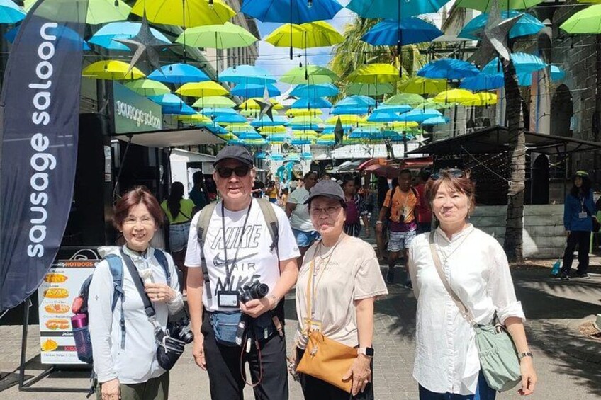 Une rue animée où l’on voit des gens se promener, un stand de saucisses et, au-dessus de leurs têtes, des parasols aux couleurs vives. 2