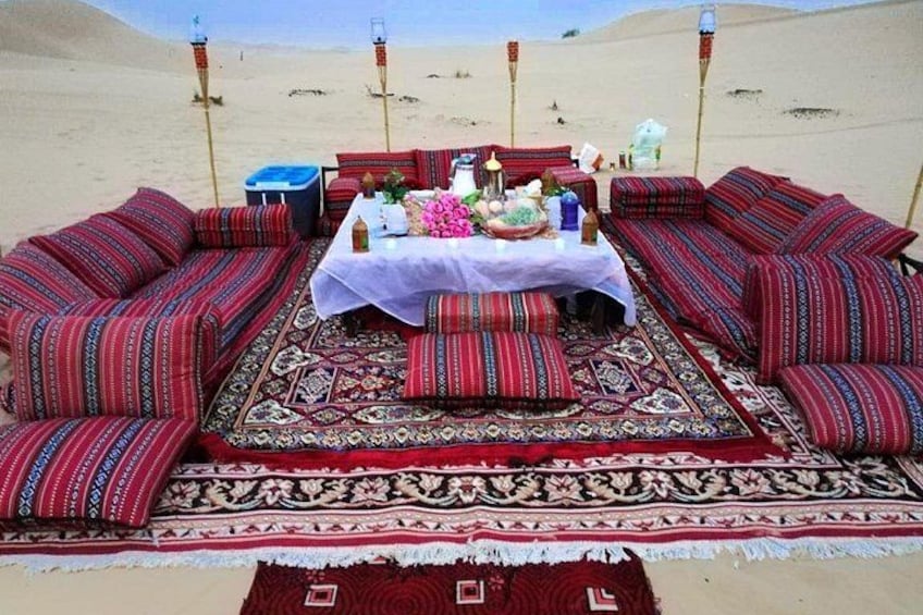 Un coin pique-nique aménagé dans le désert, avec un tapis à motifs, des coussins rayés de rouge et de noir, et une table sur laquelle sont disposés divers objets. 5