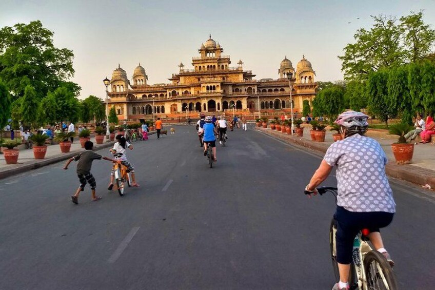 A group of people riding bicycles on a wide street with a large, ornate building in the background. 1