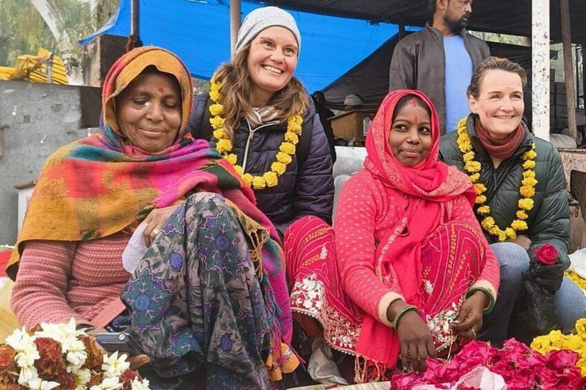 Four women sitting on a bench, adorned with garlands and traditional attire, in an outdoor setting with a blue tarp overhead. 3