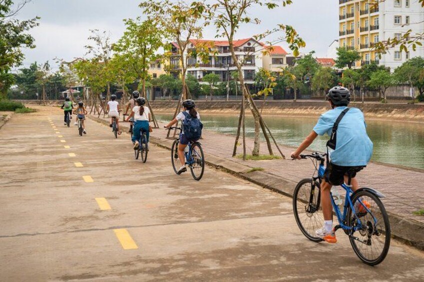 Een groep fietsers op een verhard pad langs een waterpartij, met gebouwen op de achtergrond. 2