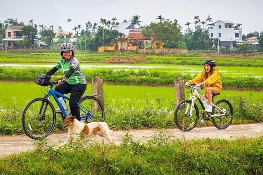 Twee fietsers op een landelijke weg, met groene velden en huizen op de achtergrond. 1