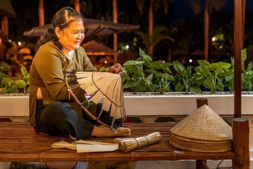 A person sitting on a wooden bench outdoors, working with a woven fan and a conical hat. 1