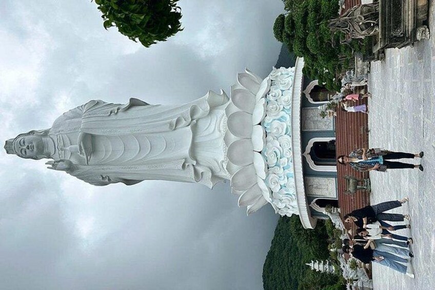 A large white statue of a figure with a lotus base, surrounded by greenery and a paved area with people. 2