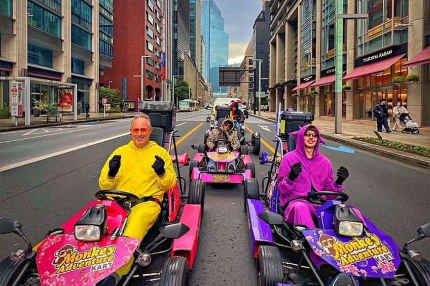Two individuals in bright yellow and purple costumes driving toy cars on a city street. 5