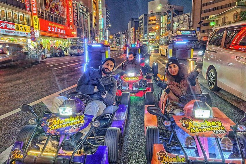 Drei Personen auf einer beleuchteten Go-Kart-Bahn in einer Stadtlandschaft mit Gebäuden und Fahrzeugen im Hintergrund. 3