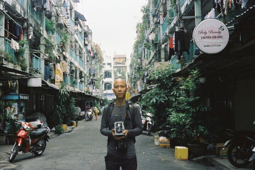 A narrow street with buildings, a person holding a camera, and a red motorcycle. 1