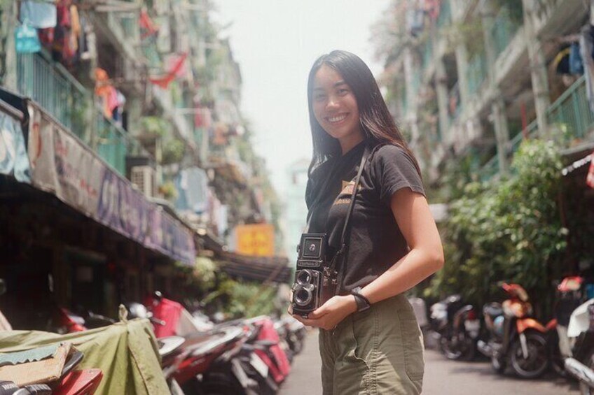A person holding a camera on a street with buildings and parked motorcycles. 2