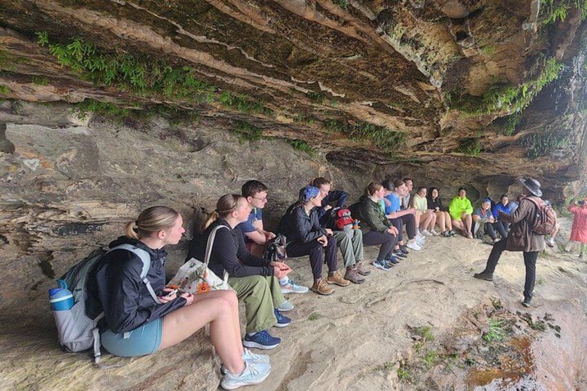A group of people sitting on a rocky ledge, some with backpacks, in a natural setting with a cave-like rock formation in the background. 4