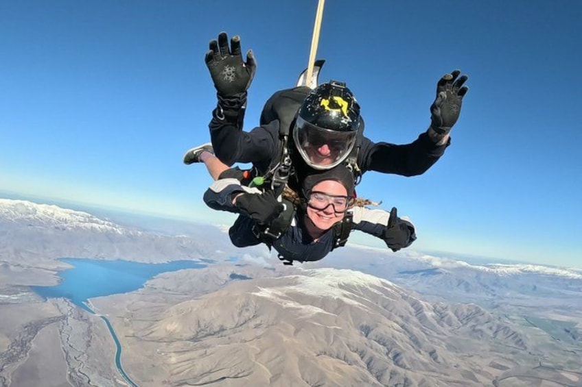 A person skydiving with a parachute, wearing a helmet and gloves, above a mountainous landscape with a river. 1
