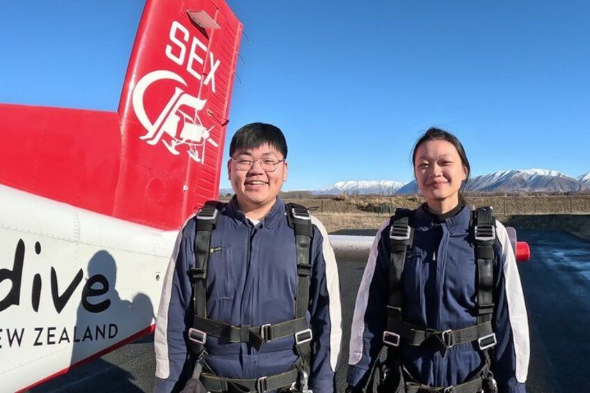 Two people standing in front of a red and white airplane with 'SEX Dive New Zealand' written on the tail. 2