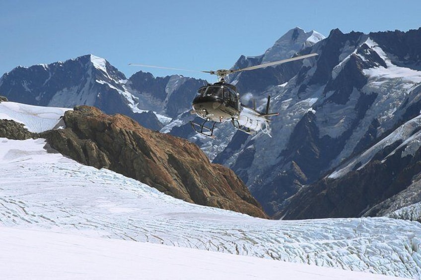 Ein Hubschrauber fliegt über eine verschneite Berglandschaft mit einem Gletscher und felsigem Gelände. 2