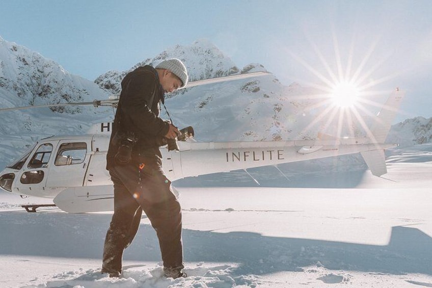 Eine Person macht ein Foto mit einer Kamera, stehend vor einem mit 'INFLITE' beschrifteten Helikopter auf einer verschneiten Fläche mit Bergen im Hintergrund. 4