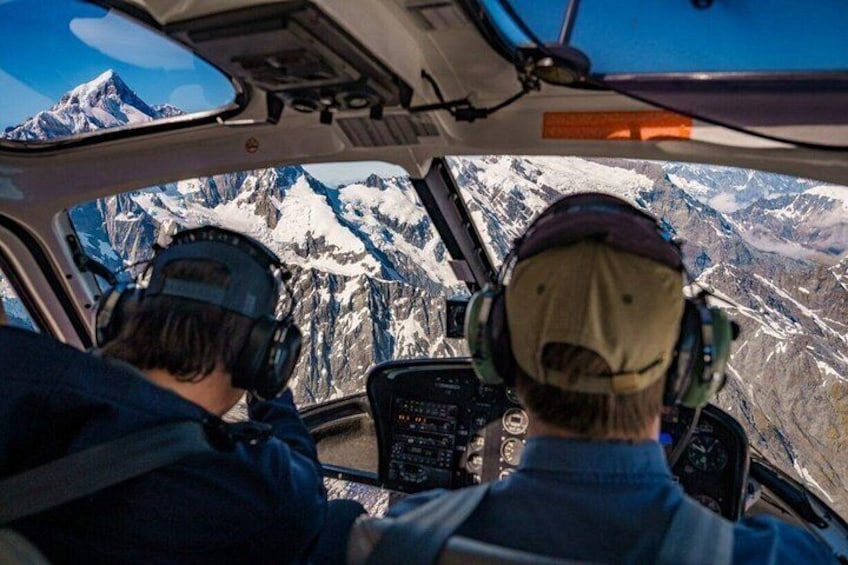Ein Hubschrauber-Cockpit mit zwei Piloten, eine gebirgige Landschaft und klarer Himmel. 5