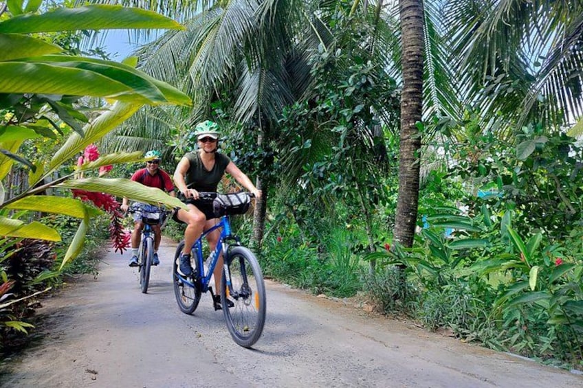 Two cyclists on a paved road surrounded by lush greenery and palm trees. 2