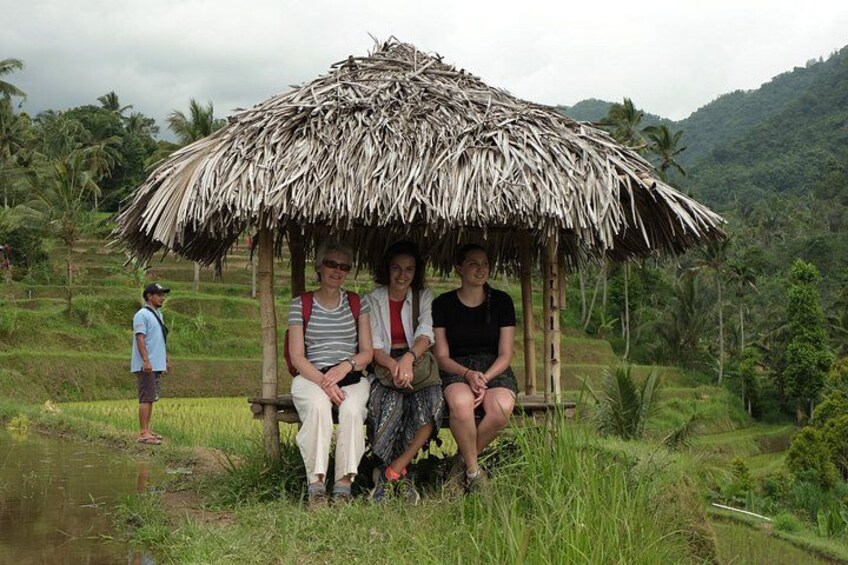 A group of people sitting under a thatched structure in a rural setting with rice paddies and palm trees in the background. 5
