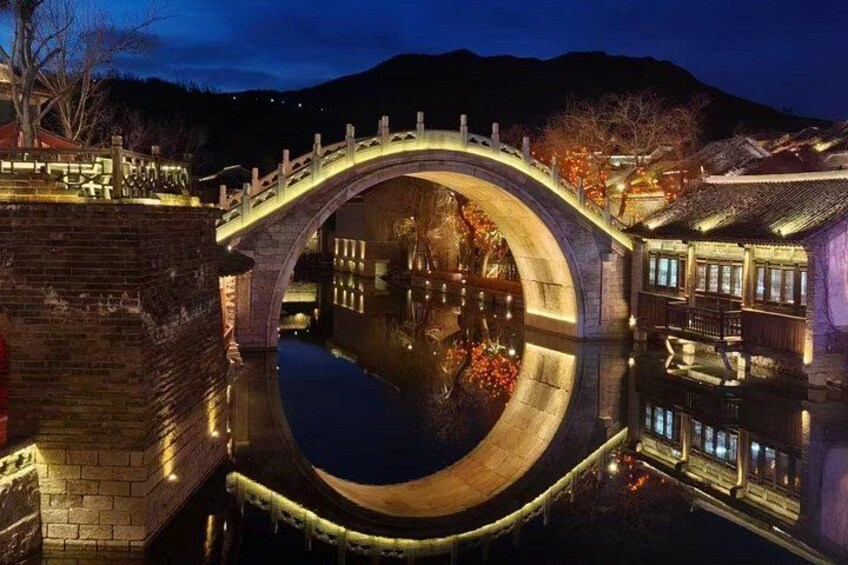 A stone bridge over a calm river at night with illuminated buildings on either side. 4