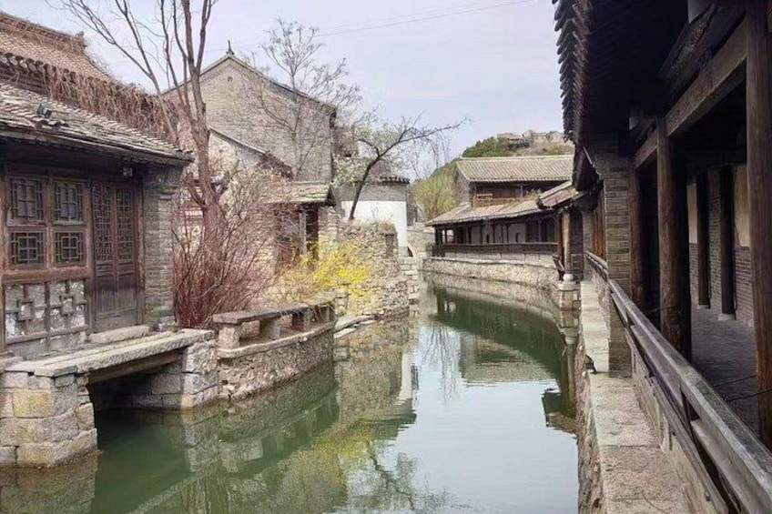 A traditional Chinese courtyard with stone buildings, a narrow water canal, and a wooden bridge. 1